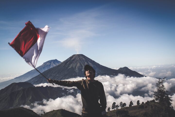 man wearing black crew neck sweater holding white and red flag standing near mountain under blue and white sky